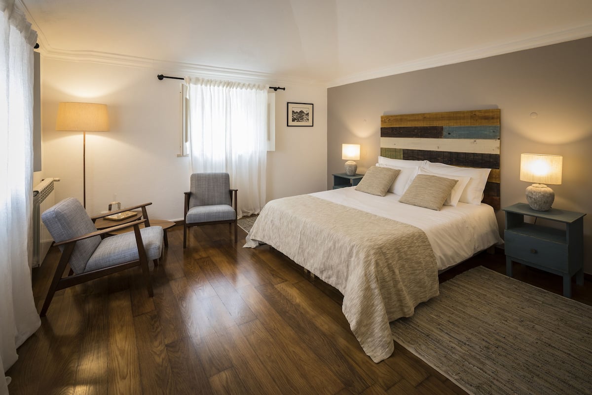 Quarto de hotel com uma cama grande, duas mesas de cabeceira com luminárias, uma cadeira, janela com cortinas e um quadro na parede.
