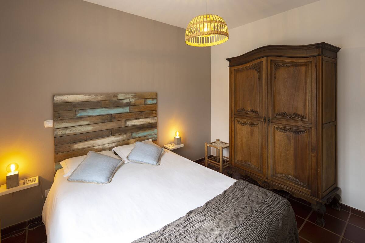Quarto com cabeceira de madeira, cama com roupa de cama branca, mesa de cabeceira com abajur e guarda-roupa de madeira.