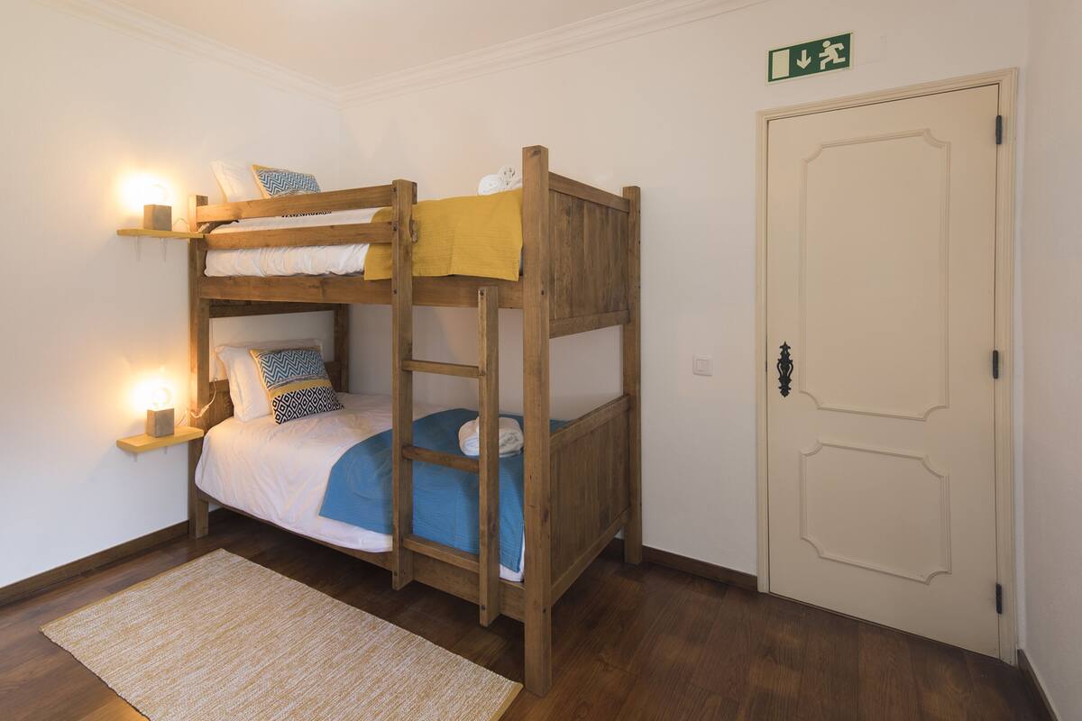 Quarto com beliche, cabeceira de madeira, mesa de cabeceira com abajur e porta com placa de saída.