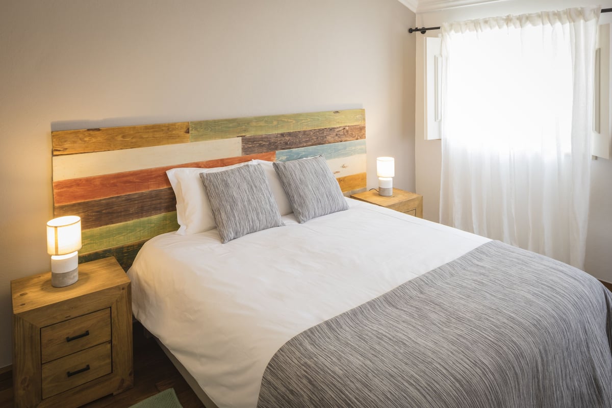 Cama bem arrumada com cabeceira de madeira, duas luminárias de mesa e mesa de cabeceira com gavetas.