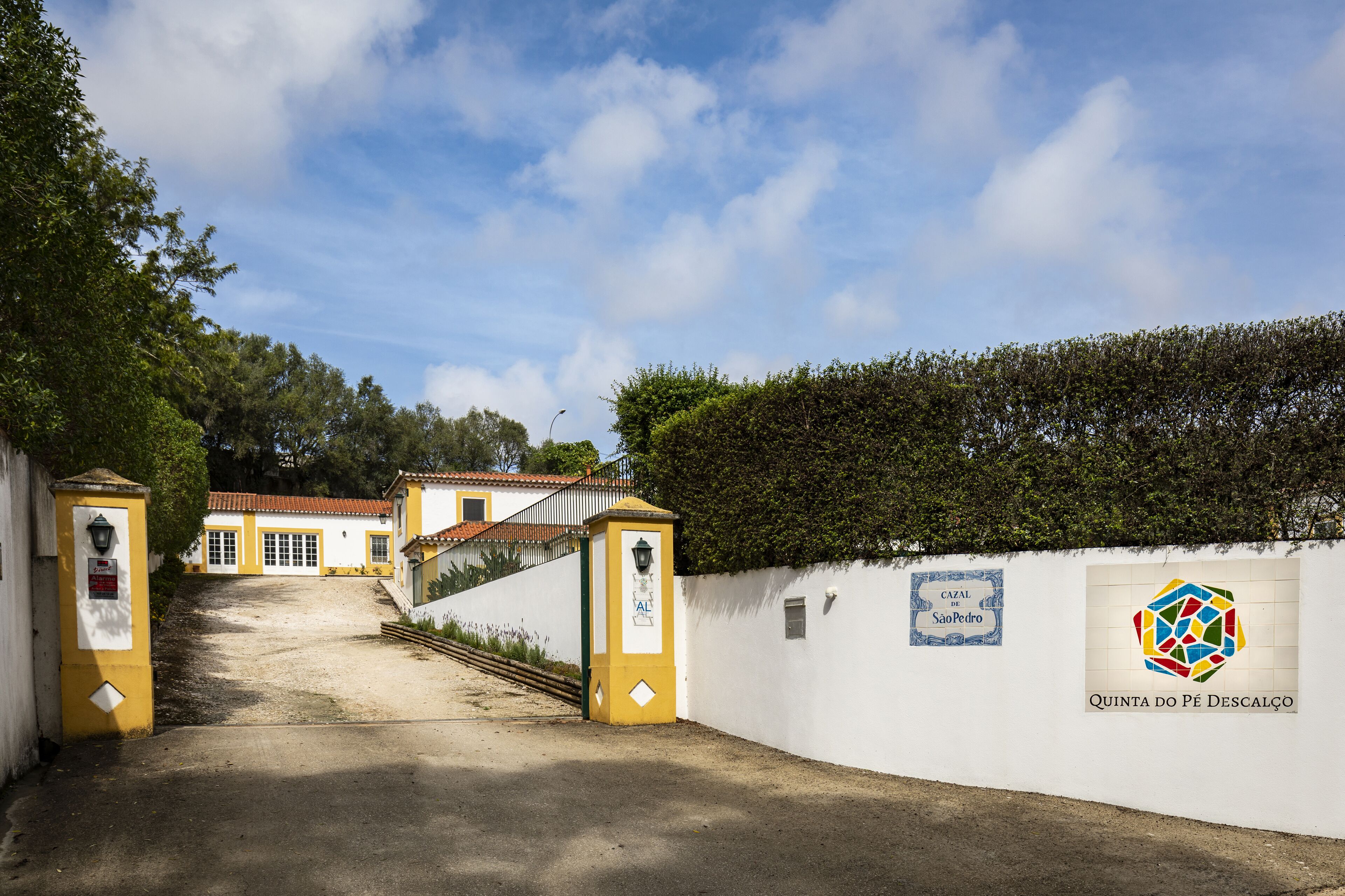 A pathway leading to a building with a white facade and yellow accents, a sign reading "Quinta do Pé Descalço", and a logo with a colorful geometric design.