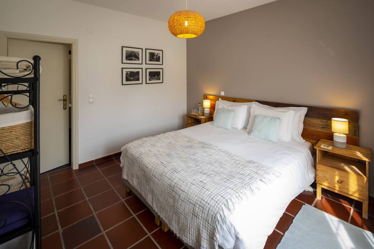 Quarto bem arrumado com uma cama grande, duas mesas de cabeceira com abajures, uma cabeceira de madeira e uma porta que dá para outro cômodo.