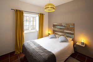 A neatly made bed with a wooden headboard, two bedside lamps, and a window with yellow curtains. - Quinta do Pé Descalço Guesthouse Sintra (Sintra)
