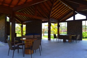 Un patio techado al aire libre con pilares de piedra, vigas de madera y varias mesas y sillas.