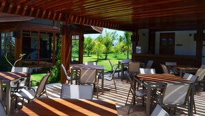 Un patio al aire libre con mesas y sillas de madera, una zona cubierta con techo de madera y vistas a un jardín bien cuidado con palmeras.