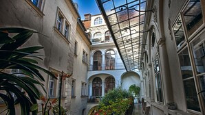 Une ruelle étroite, bordée de fenêtres en arc de cercle et de balcons, et entourée d’une végétation luxuriante et de plantes en pot.