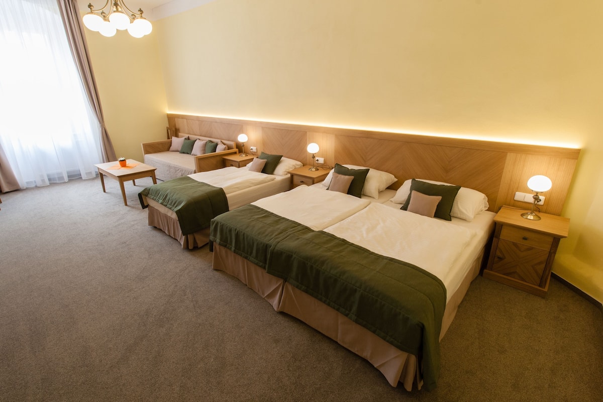 A hotel room with three beds, a small table, bedside lamps, and a large window with curtains.
