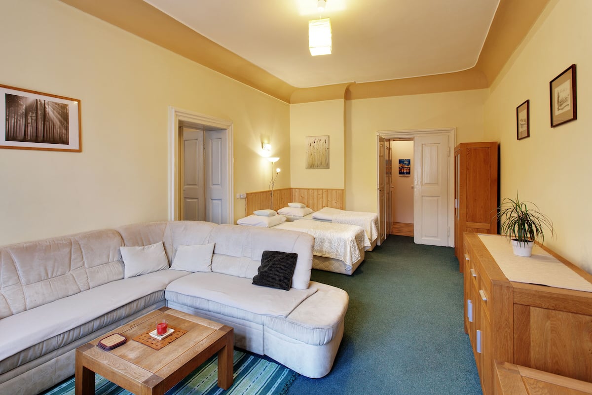 A room with a large L-shaped sofa, two single beds, a wooden cabinet, and a small potted plant.