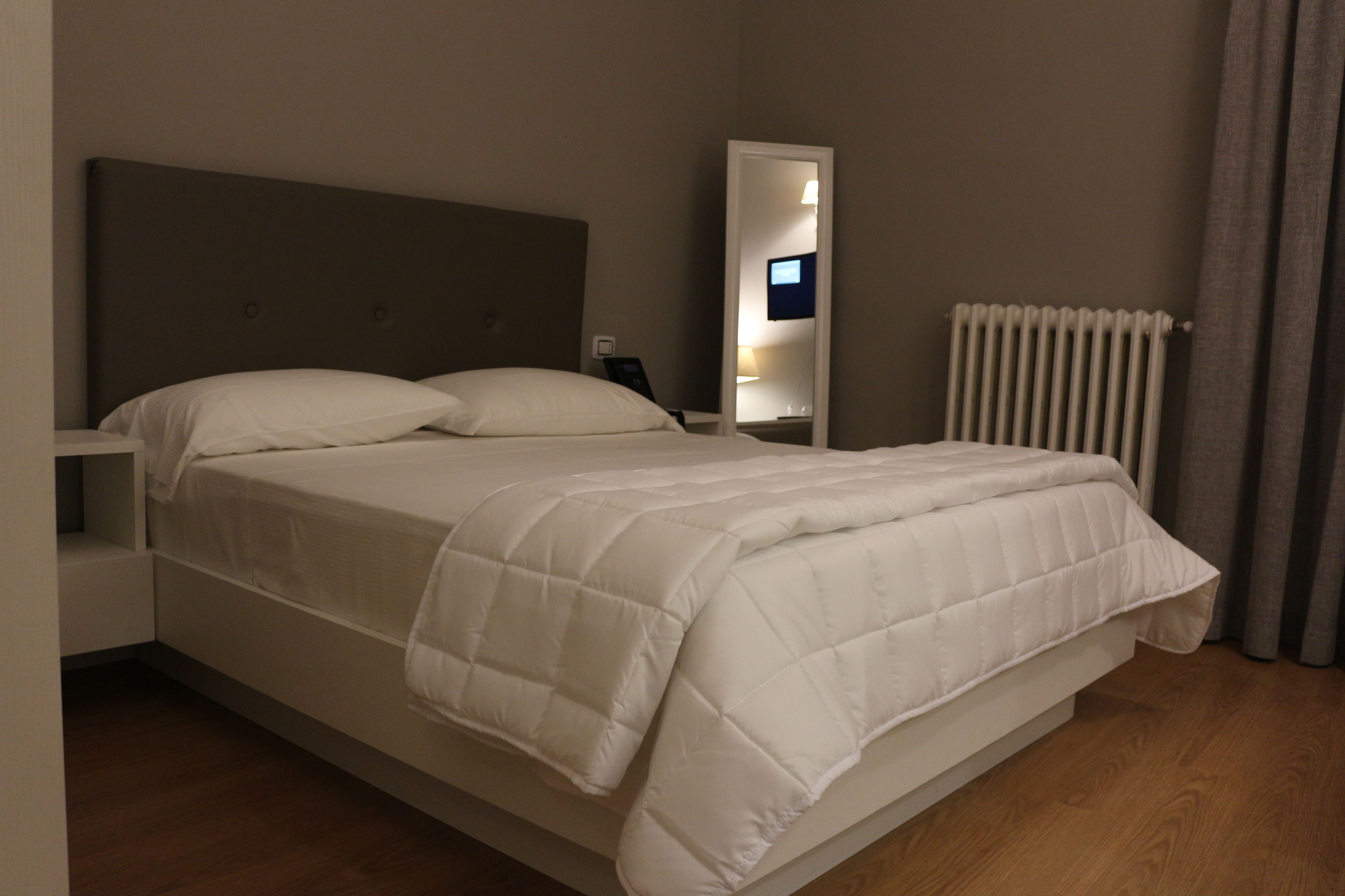 Una cama bien tendida con un edredón blanco y almohadas, una mesita de noche con una lámpara, un espejo y un radiador.