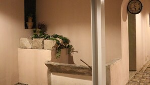 Een smalle gang met stenen vloer, een klein raam met een bloempot en een klok aan de muur.