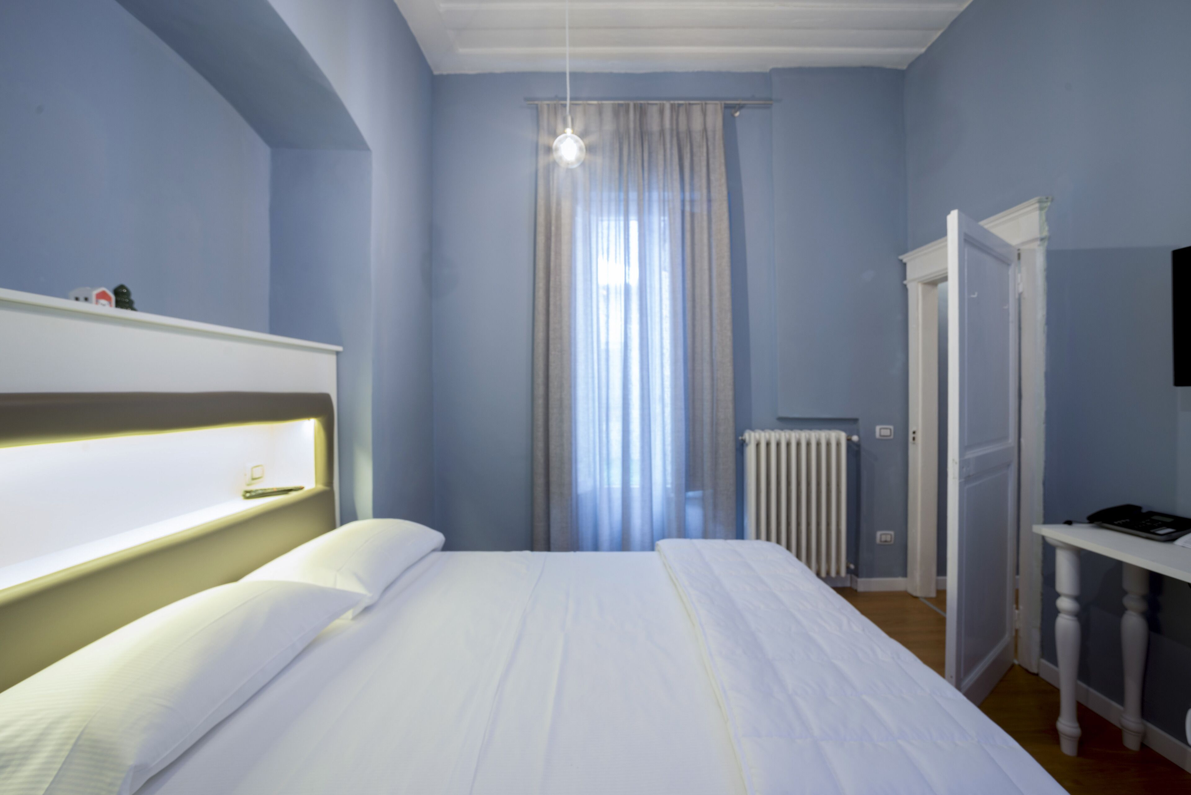 Un dormitorio con cama, mesita de noche, radiador y ventana con cortinas.