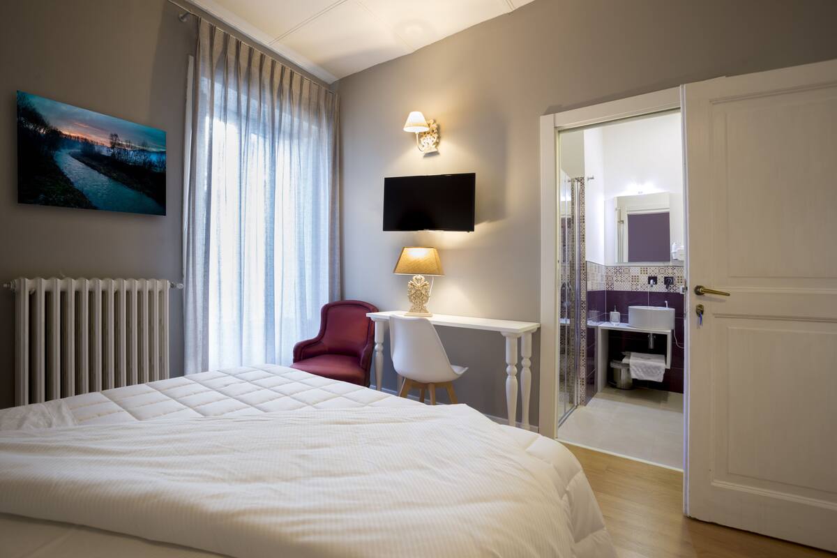 A hotel room with a large bed, a desk with a lamp, a TV mounted on the wall, and a bathroom visible through an open door.