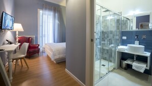 Hotelkamer met een bed, een bureau met stoel, een rode fauteuil, een douche met glazen wanden en een badkamer met wastafel en spiegel.