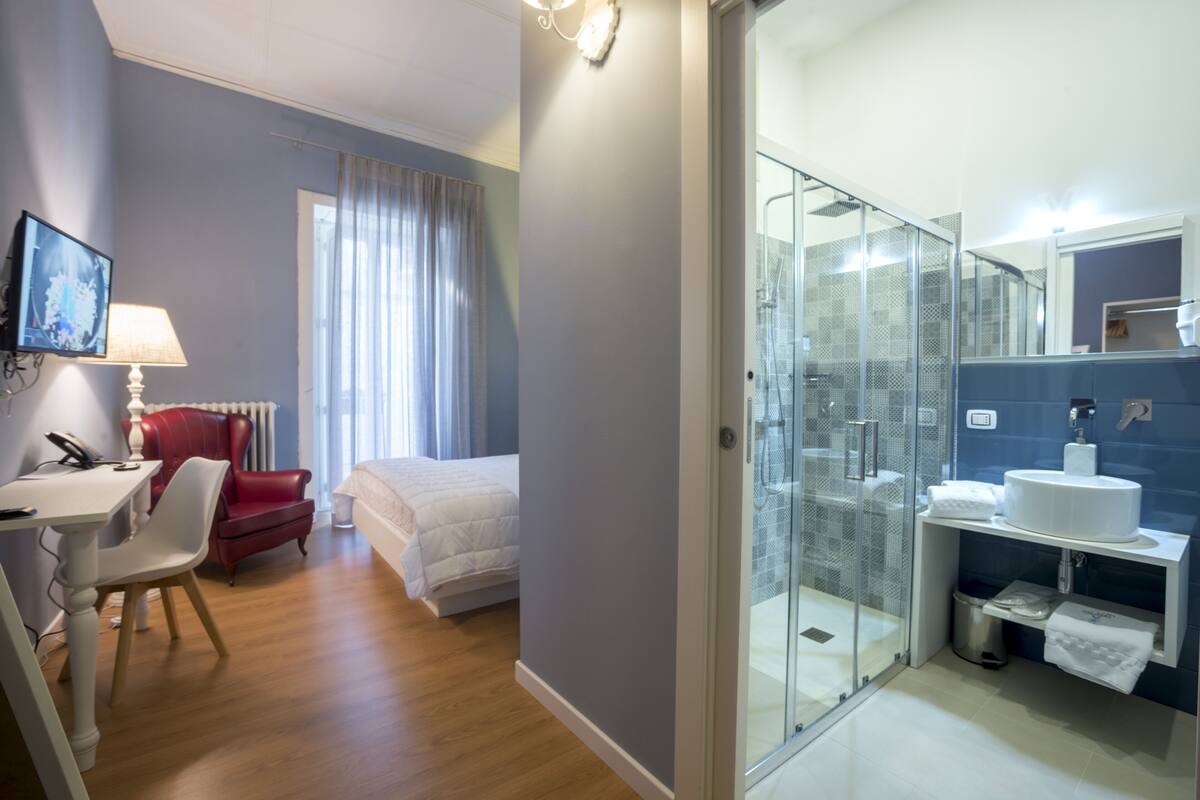 A hotel room with a bed, a desk with a chair, a red armchair, a glass-enclosed shower, and a bathroom with a sink and mirror.