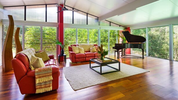 Ein geräumiges Wohnzimmer mit einem roten Sofa, einem schwarzen Klavier und großen Fenstern mit Blick auf Grünflächen.