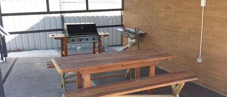 An outdoor patio with a wooden table and bench, a grill, and a sink.