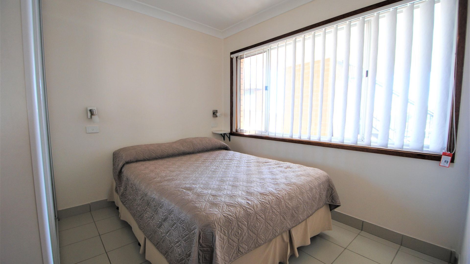 Un dormitorio pequeño y ordenado con una cama individual, una ventana con persianas y suelo de baldosas.