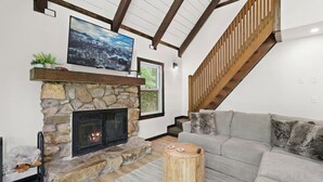 A cozy living room with a stone fireplace, a grey sofa, and a wooden staircase. - Renovated, Foosball, In the trees, EV charger! (Beech Mountain)