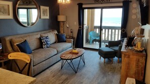 Ein gemütliches Wohnzimmer mit einer Couch, einem Couchtisch und Blick auf den Strand durch eine Schiebetür.