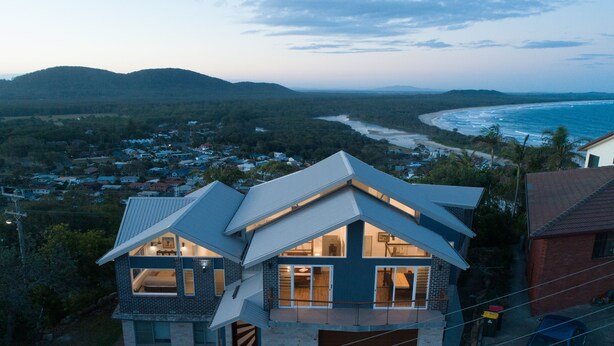 A modern house with a sloped roof, large windows, and a garage. It is surrounded by a well-maintained lawn and a view of the ocean and mountains in the background.