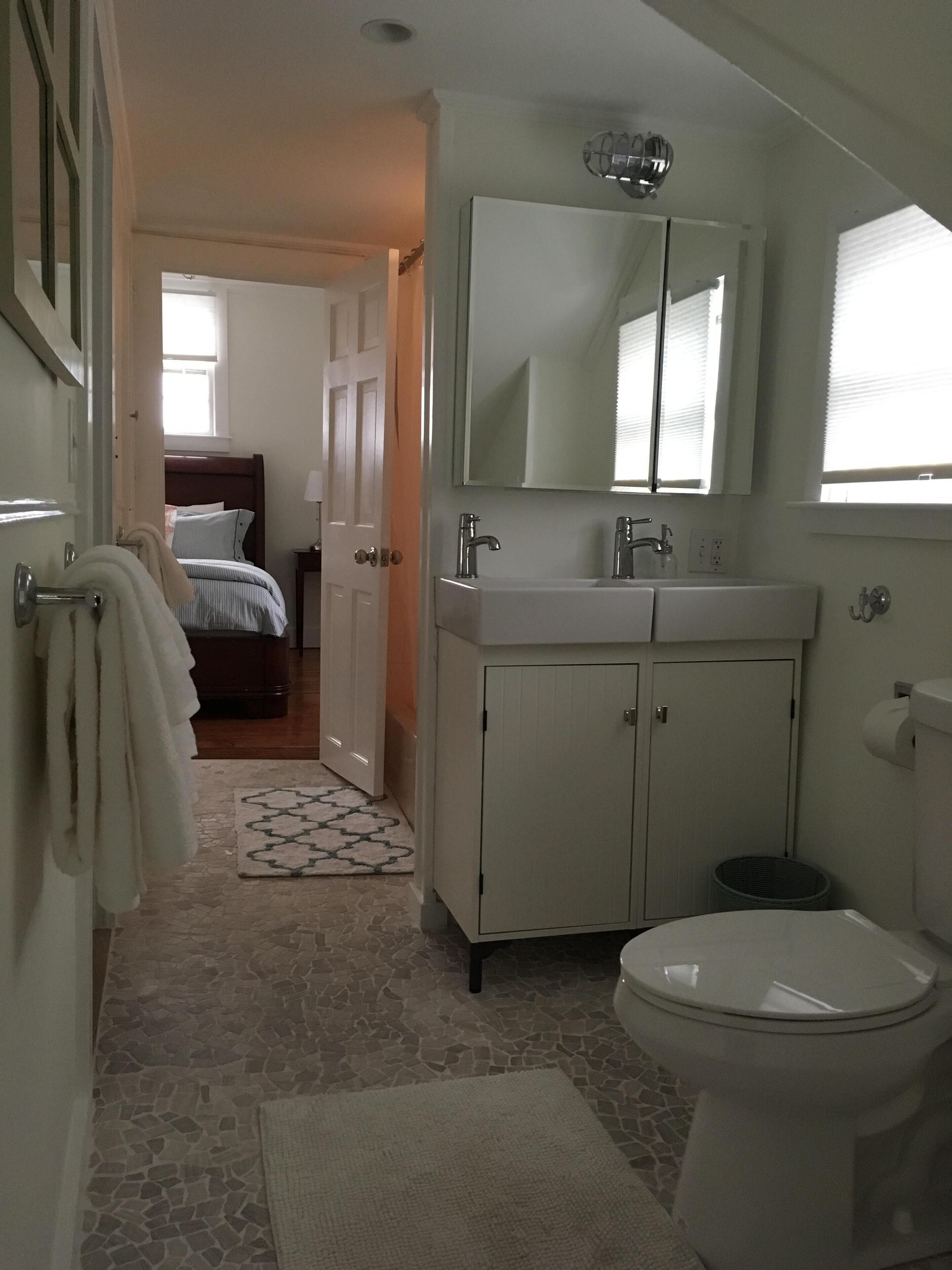 A bathroom with a toilet, sink, and mirror. There is a patterned rug on the floor and a door leading to a bedroom.