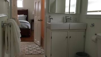 A bathroom with a toilet, sink, and mirror. There is a patterned rug on the floor and a door leading to a bedroom.