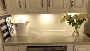 Una cocina con armarios blancos, un refrigerador de vinos empotrado con iluminación azul, un cubo blanco y un florero con flores blancas.