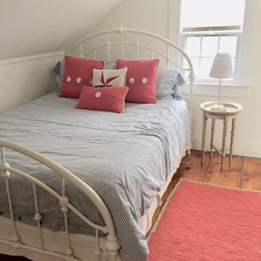 A bed with a white metal frame, a grey and white striped bedspread, and multiple pillows in shades of pink and grey. There is a small round table with a lamp next to the bed.