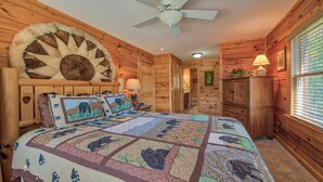 A cozy log cabin bedroom with a large bed, a ceiling fan, wooden walls, and a window with blinds. - SPECIAL RATES OCT 12-16, MT VIEWS, ATTACHED GARAGE, FLAT PAVED PARKING, PET FEE. (Maggie Valley)