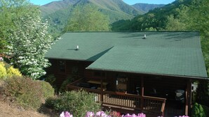 A log cabin with a green roof, surrounded by lush greenery and mountains in the background. - SPECIAL RATES OCT 12-16, MT VIEWS, ATTACHED GARAGE, FLAT PAVED PARKING, PET FEE. (Maggie Valley)
