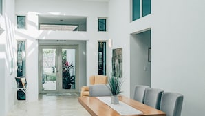 Un comedor moderno con una mesa de madera, sillas grises y un banco. Un tragaluz proporciona luz natural y el espacio interior es limpio y blanco, con vistas al exterior.