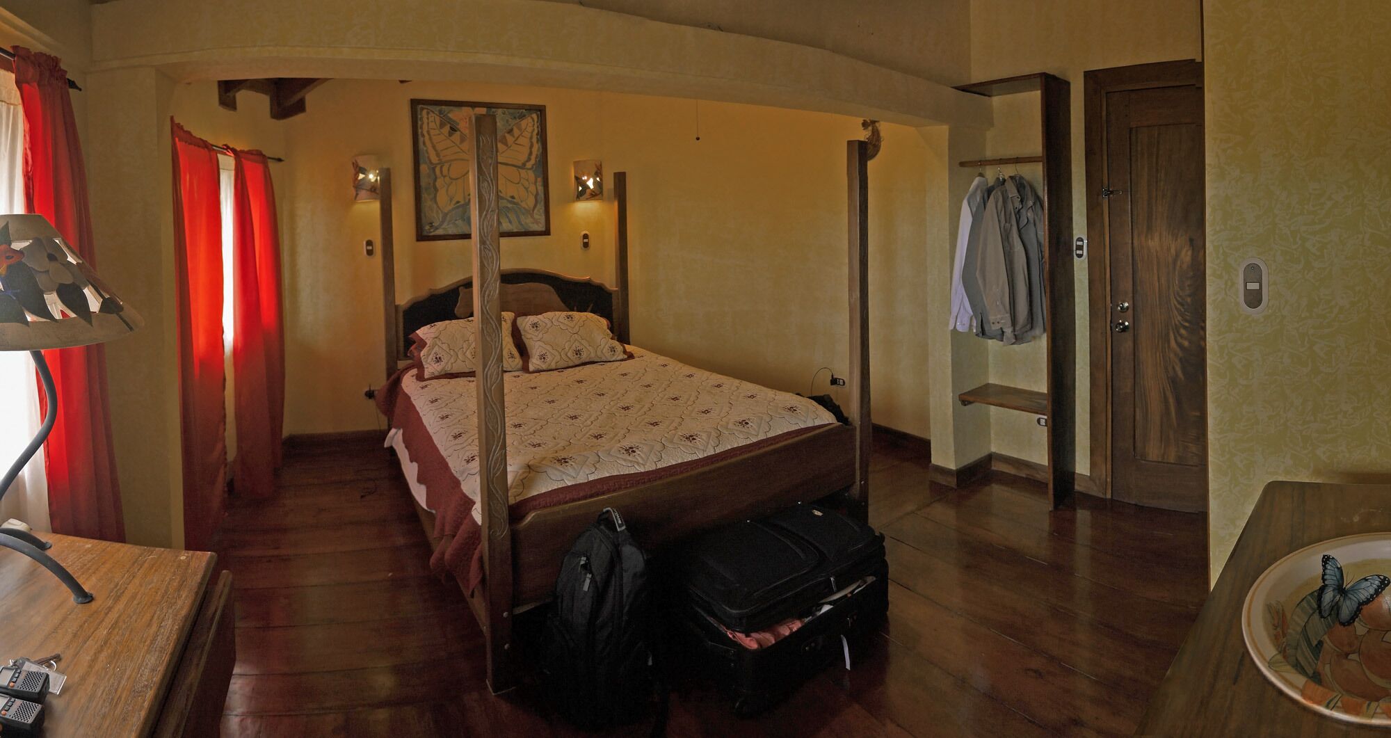 Un letto a baldacchino con copriletto floreale, un comodino in legno con una lampada, una valigia sul pavimento e un appendiabiti con abiti.