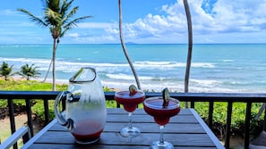 Une table avec une carafe et deux verres remplis d’une boisson rouge, donnant sur une plage bordée de palmiers et sur l’océan.