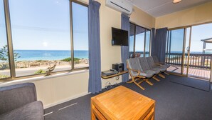 A room with a large window overlooking a beach, a flat-screen TV mounted on the wall, and a seating area with a wooden table. - Sandy Feet - 31 Gold Coast Drive (CARRICKALINGA)