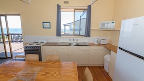 A compact kitchen with a stove, sink, and a refrigerator. There is a dining table with chairs, a view of the sea through a window, and a small shelf with a microwave. - Sandy Feet - 31 Gold Coast Drive (CARRICKALINGA)