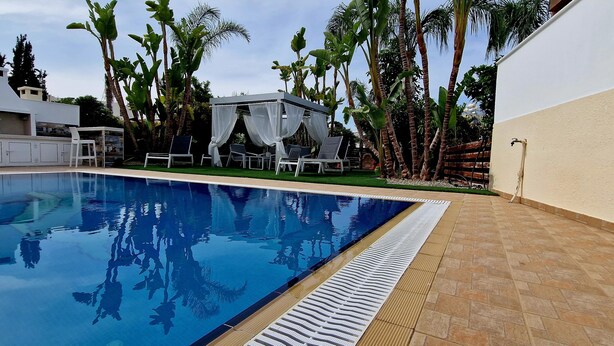 A swimming pool with lounge chairs and a covered seating area, surrounded by palm trees and a white building. - Villa With Private Pool, Sea View With Grass And 2 Gazebos (Pernera)