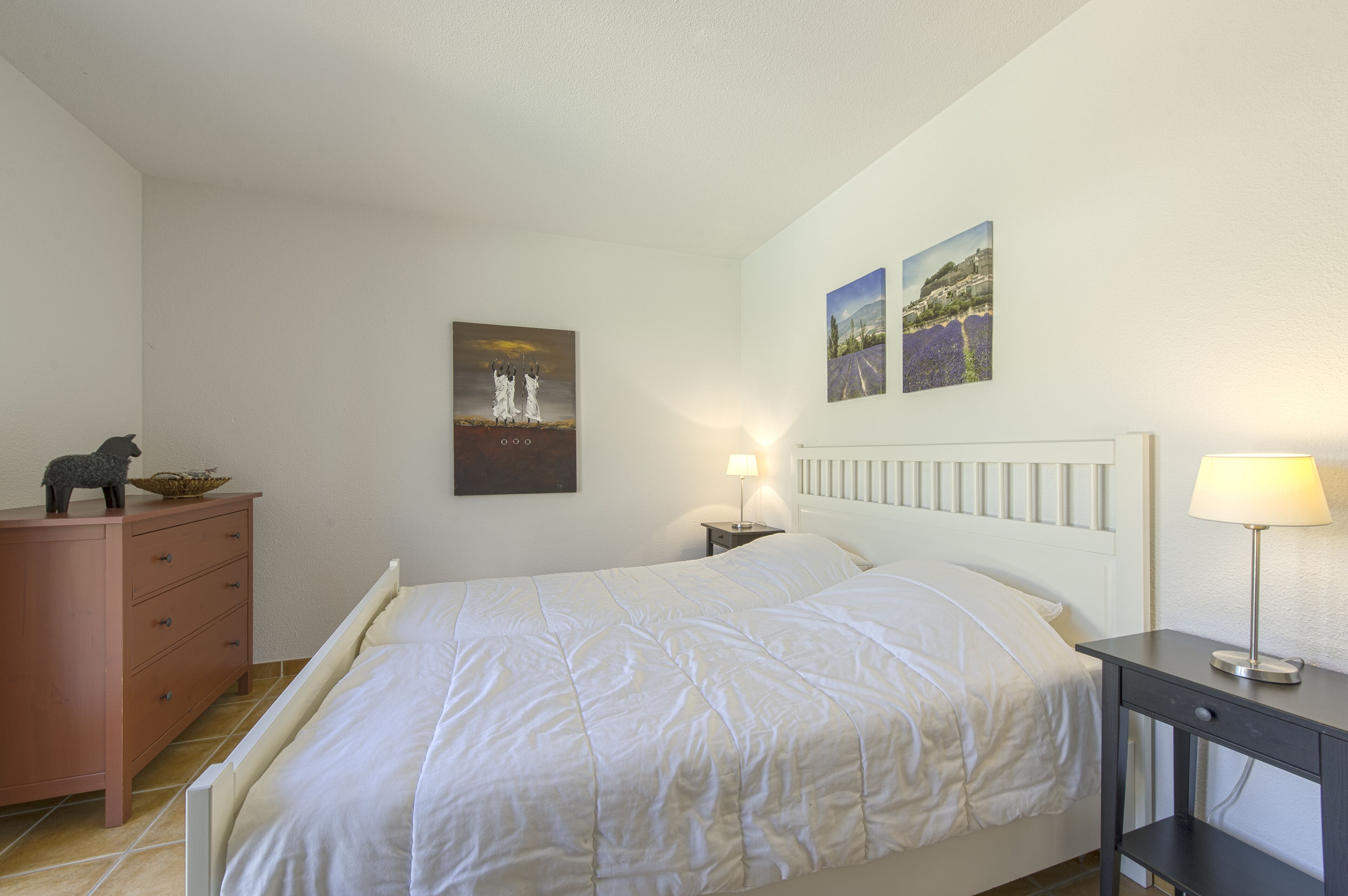A bedroom with a bed, a dresser, a nightstand with a lamp, and two wall-mounted paintings.