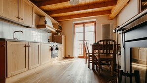 A kitchen with wooden cabinets, a stove, and a dining area with a chair. - Cozy Cottage on the mountains in Trentino Alto Adige Valle dei Mocheni (SANT'ORSOLA TERME)