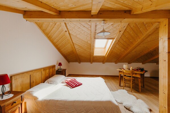 Un acogedor dormitorio en el ático con techo de madera, un tragaluz, una cama con ropa de cama blanca, un almohadón rojo, una mesita de noche de madera con una lámpara y una silla rústica.