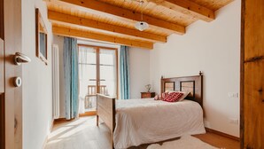 A bedroom with a wooden ceiling, a bed with white bedding, a fluffy rug, and a view of a balcony through a window.