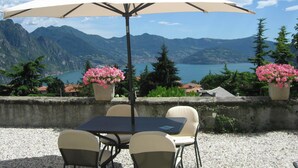An outdoor seating area with a table and chairs, a large umbrella, and two flower pots with pink flowers. - PANORAMA Apartment - Large lake view apartment, 2 rooms, 4 places (Solto Collina)