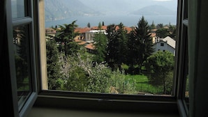 A window view of a residential area with houses, trees, and mountains in the background. - PANORAMA Apartment - Large lake view apartment, 2 rooms, 4 places (Solto Collina)