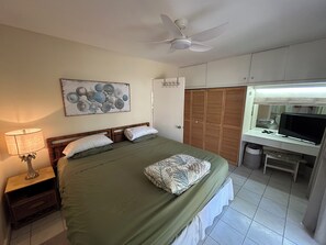 A bedroom with a bed, a nightstand, a lamp, a ceiling fan, a wardrobe, a desk with a computer, and a wall-mounted TV.
