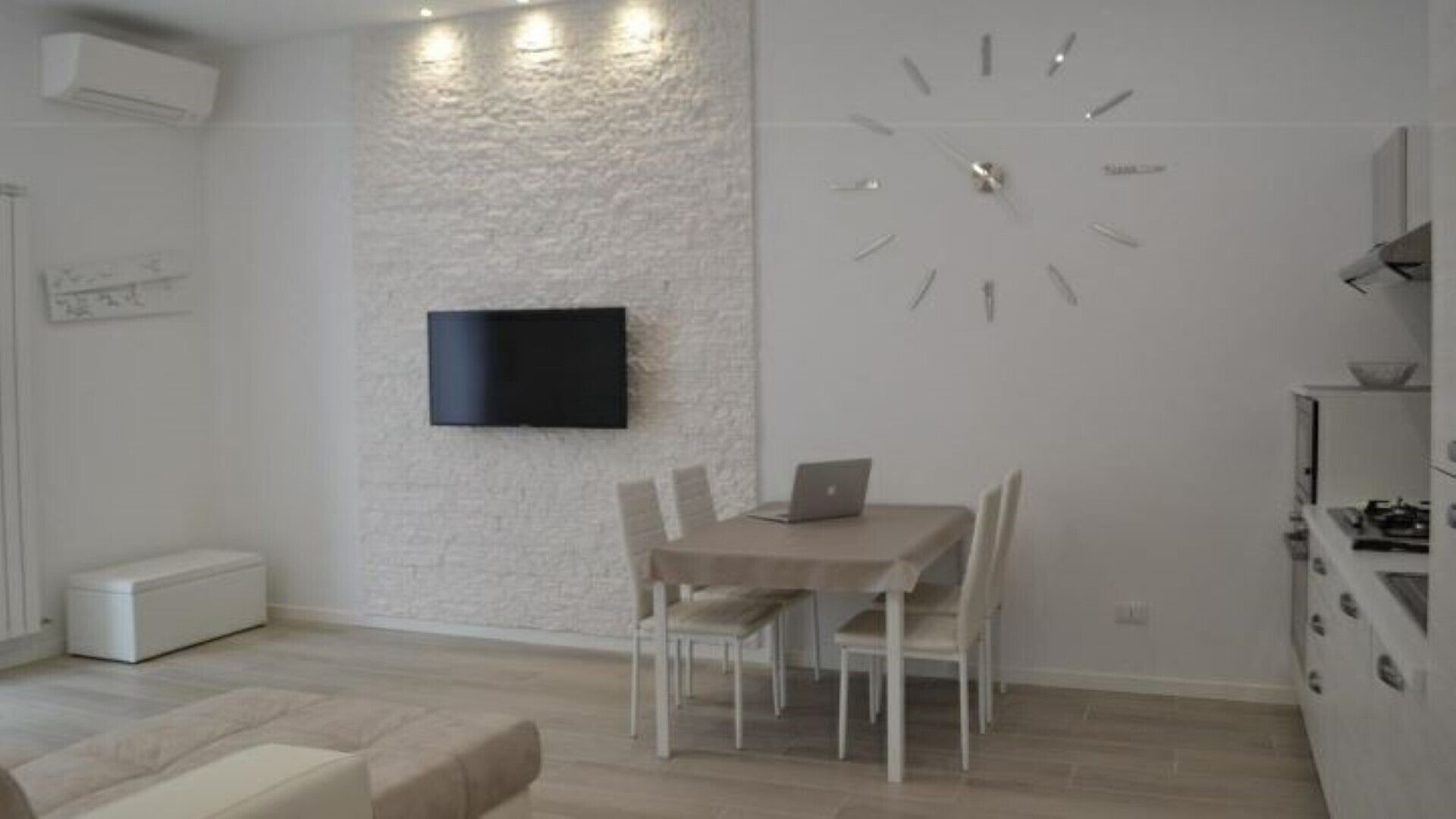 A modern kitchen with a dining area, a flat-screen TV mounted on the wall, and a large wall clock.