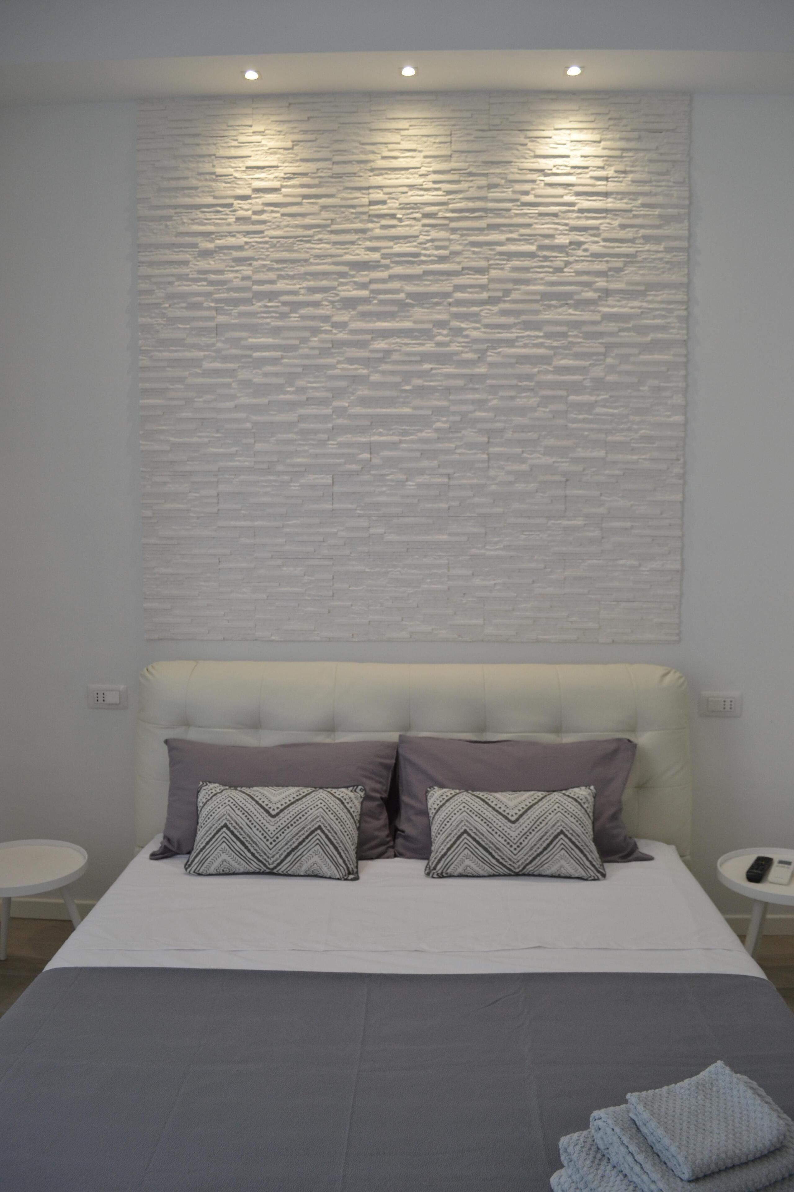 Une chambre à coucher moderne avec une tête de lit blanche à motifs de briques, deux oreillers, une couvre-lit grise et une petite table de chevet avec une télécommande.