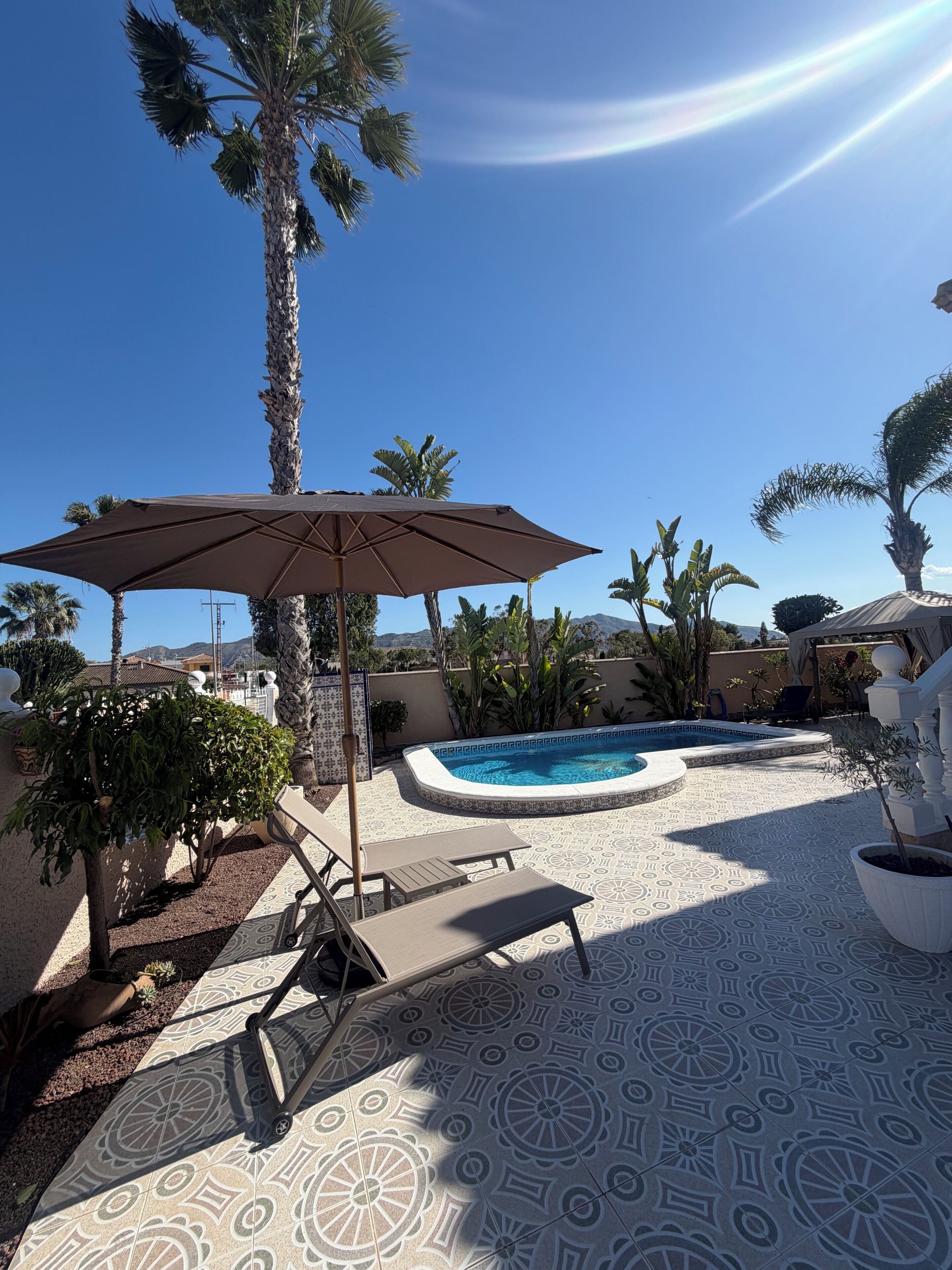 A pool area with a patterned floor, lounge chairs, and a palm tree.
