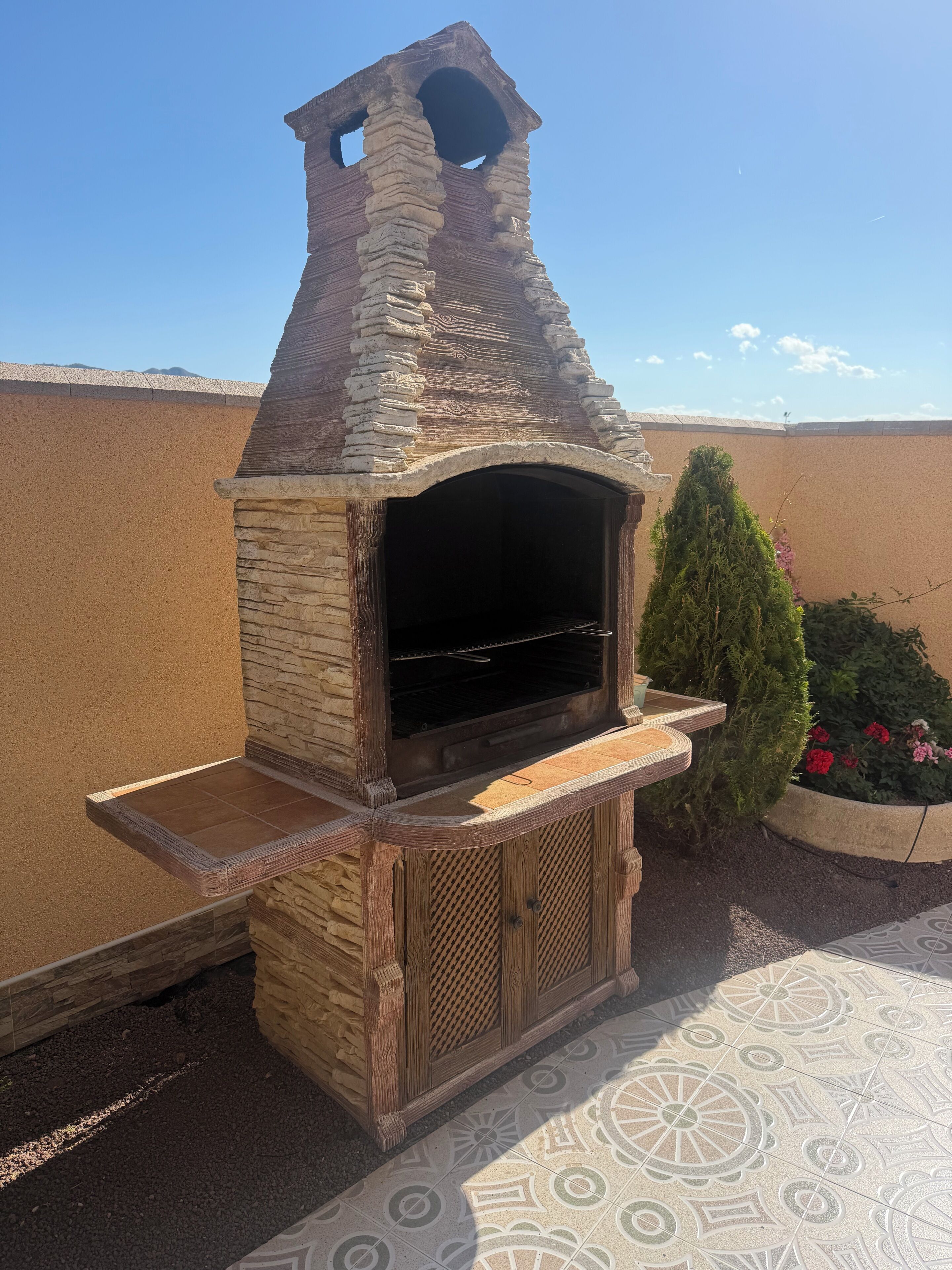 Uma churrasqueira de pedra com chaminé, bancada de madeira e chão decorado com azulejos.