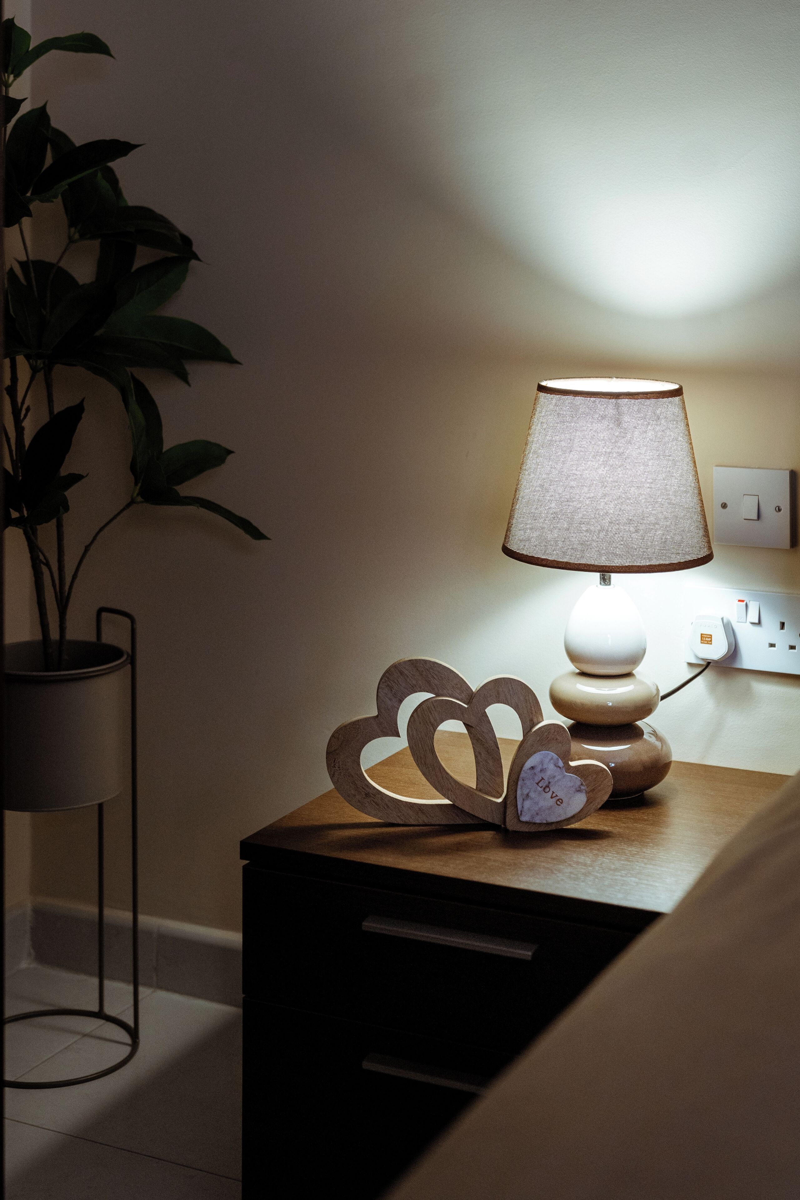 Une table de chevet avec une lampe, un objet décoratif en forme de cœur et une plante en pot.