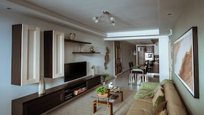 En moderne stue med et stort fladskærms-tv, en mørk træ-tv-møbel, en beige sofa og et spiseområde i baggrunden.