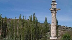 Un monument de pierre orné de sculptures complexes, entouré d’un socle en pierre et d’une clôture métallique, se dressant sur un fond de arbres et d’un paysage vallonné.